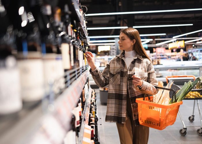 Young woman with shopping basket looking at racks with wine in store. Girl chooses wine in the supermarket